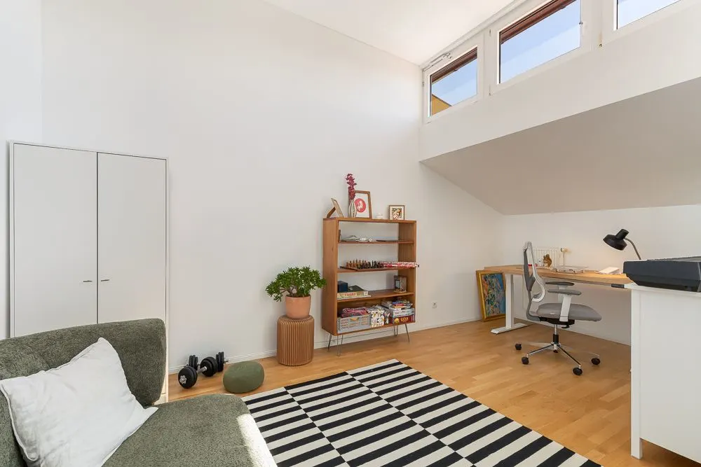 Helles Zimmer mit Schreibtisch, Regal, grünem Sofa und schwarz-weiß gestreiftem Teppich.