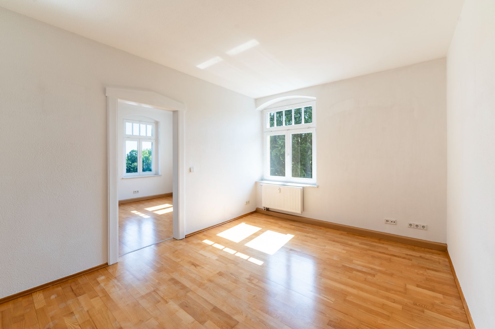 Heller, leerer Raum mit Holzboden und zwei großen Fenstern, eins davon im Nebenzimmer.