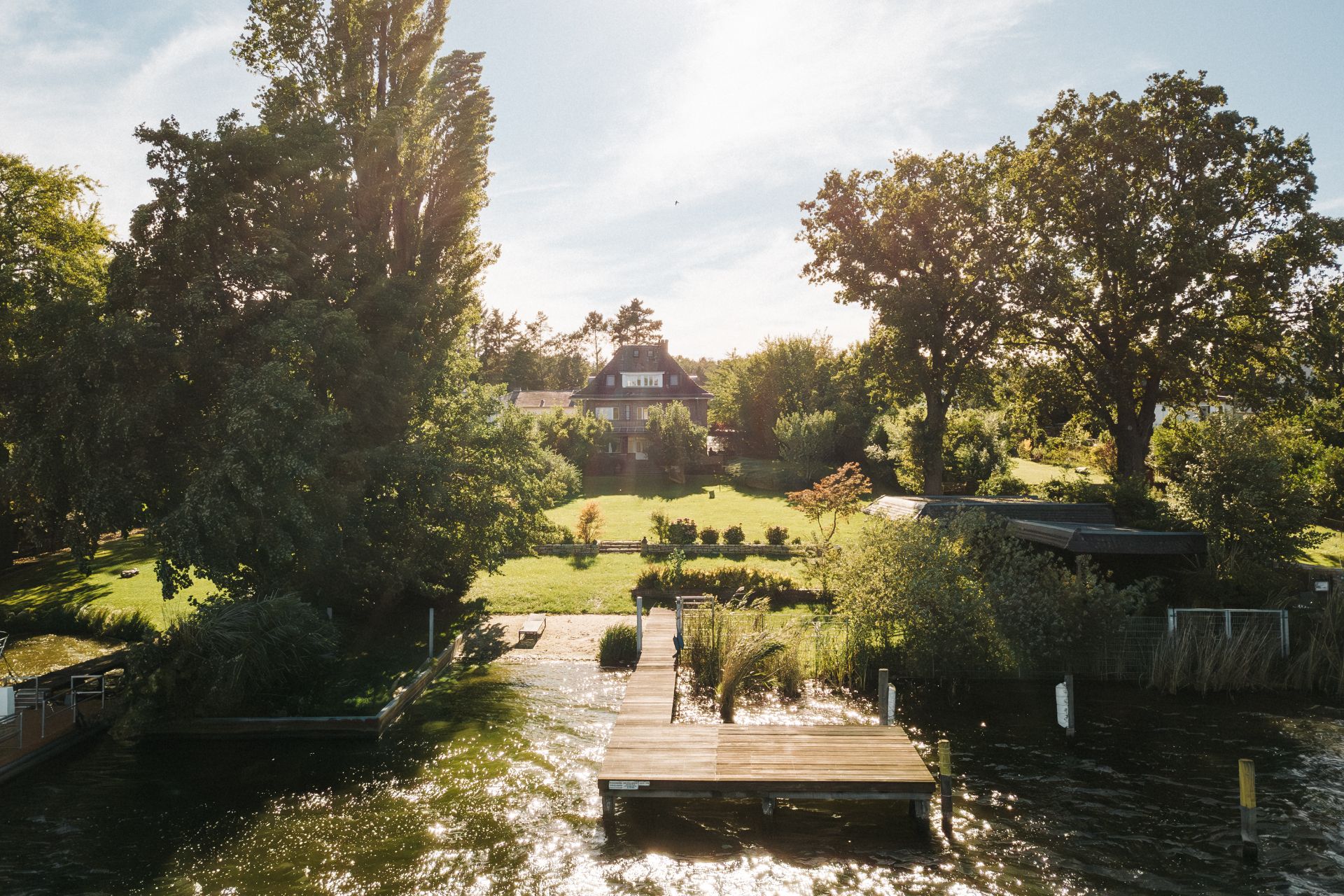 Garten mit Steg am Wasser, im Hintergrund ein Wohnhaus und große Bäume.