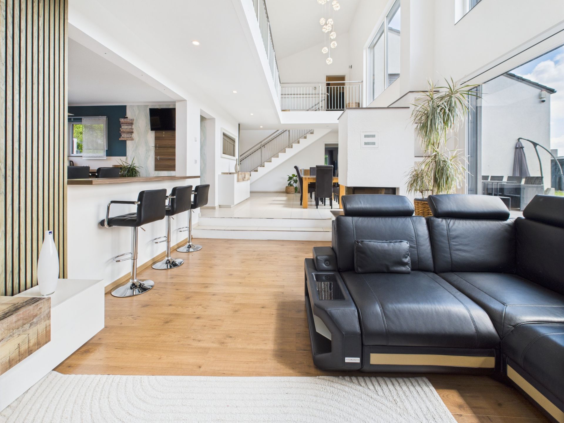 Modernes Wohnzimmer mit schwarzem Sofa, Barhockern und großer Fensterfront mit viel Tageslicht.