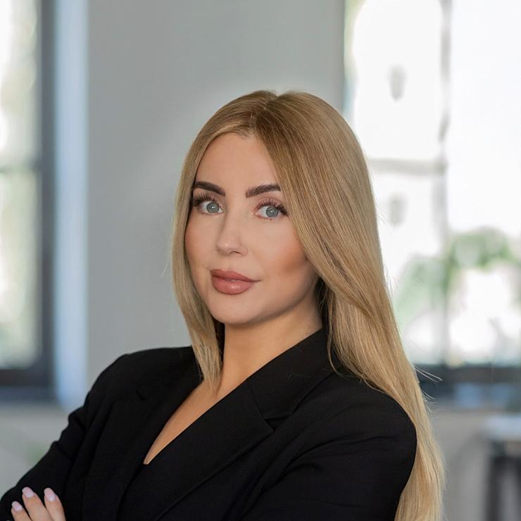 Frau mit langen blonden Haaren und schwarzem Blazer steht mit verschränkten Armen in einem Raum.