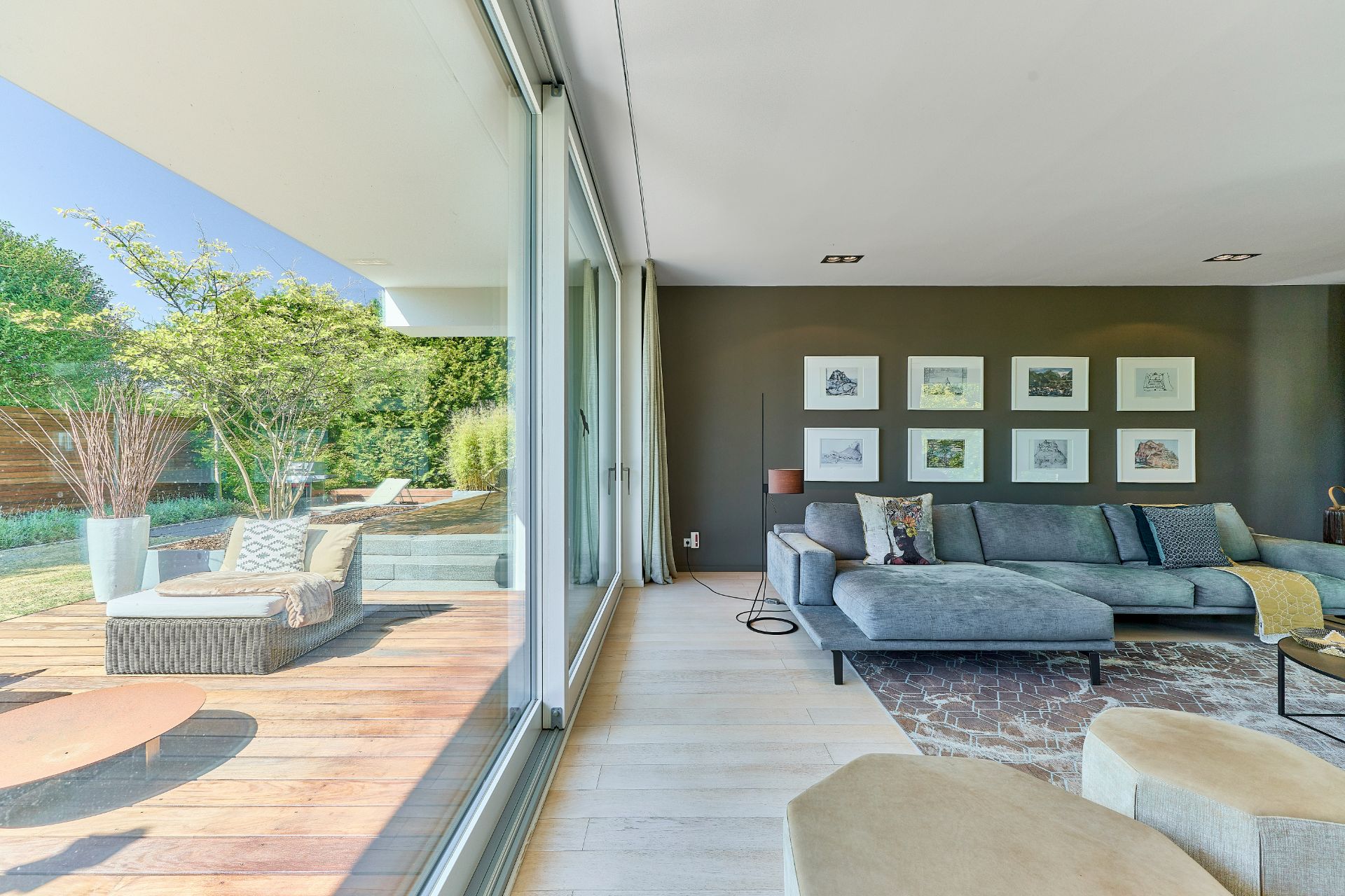 Modernes Wohnzimmer mit grauem Sofa, großer Fensterfront und Blick auf Terrasse mit Sitzmöbeln.