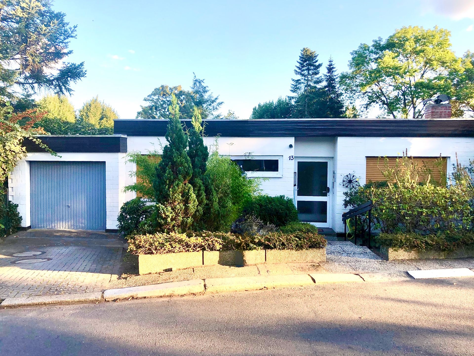 Weißes Einfamilienhaus mit Flachdach, Garage und Vorgarten mit Sträuchern.