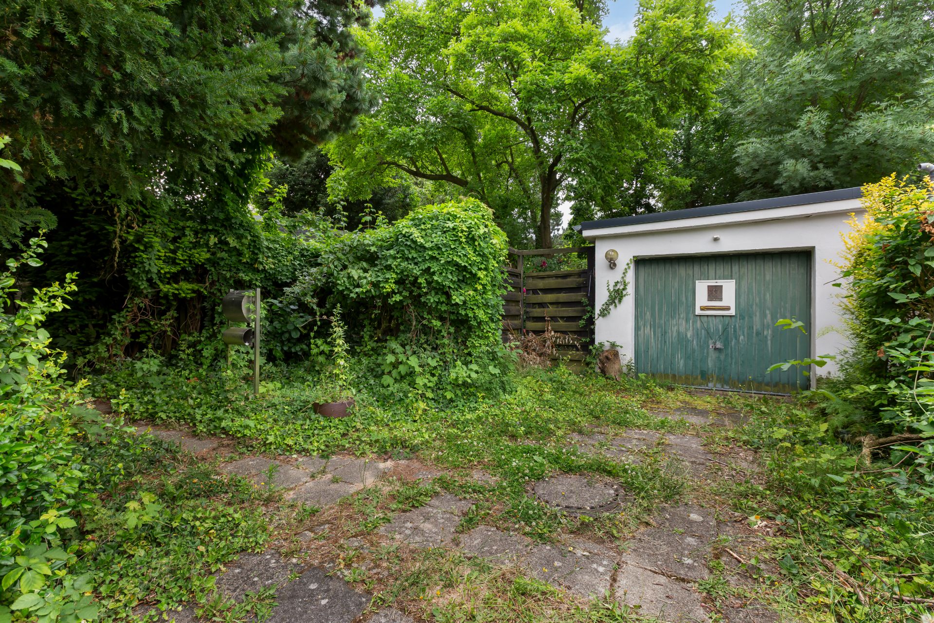 Einfahrt mit Pflastersteinen, umgeben von viel Grün, und einer Garage mit grünem Tor.