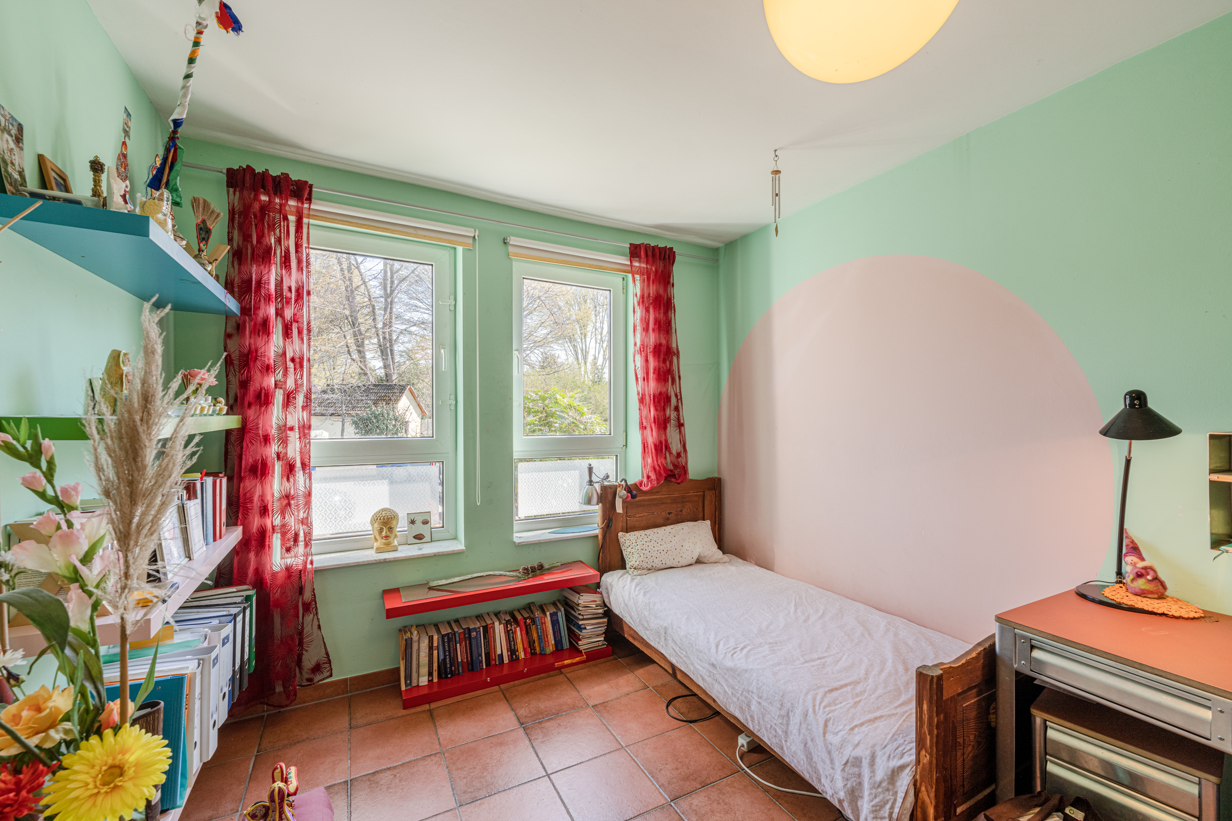 Helles Zimmer mit Einzelbett, Schreibtisch, Regal, vielen Büchern und zwei Fenstern mit roten Vorhängen.