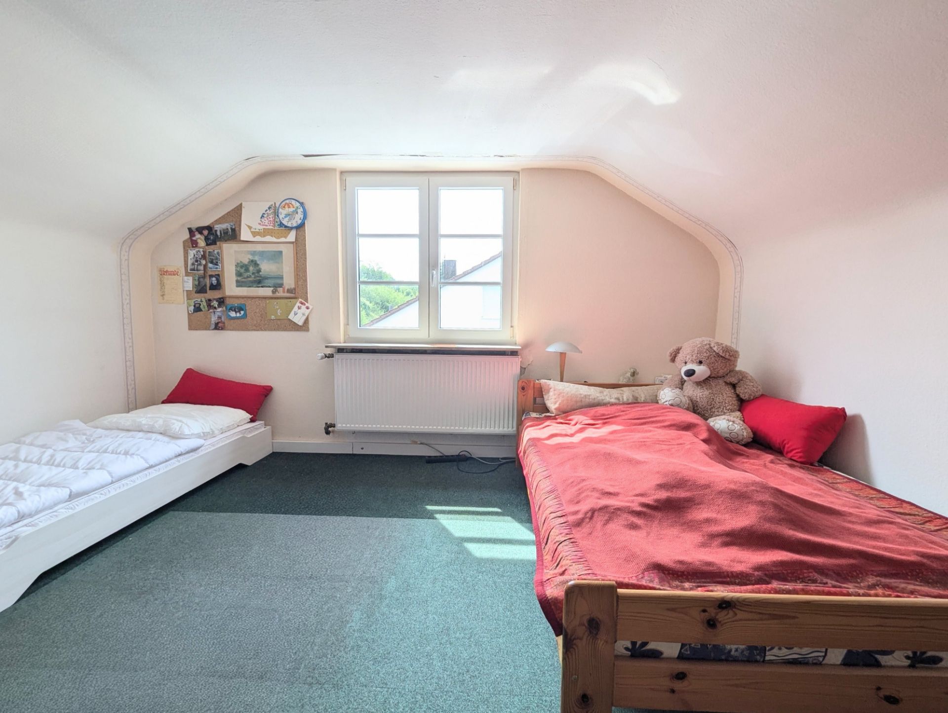 Helles Schlafzimmer mit zwei Betten, einem Fenster und einem Plüschbären auf dem rechten Bett.