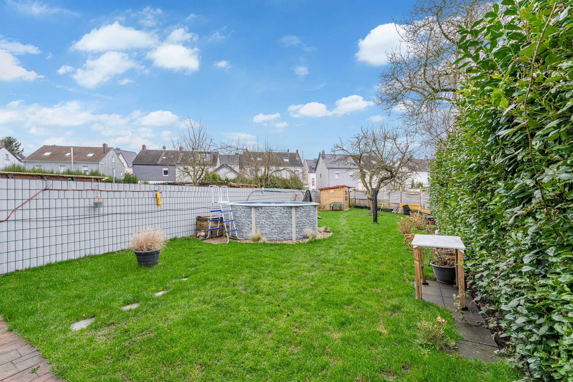 Grüner Garten mit einem runden Pool, Hecke, Bäumen und Gartenmöbeln.