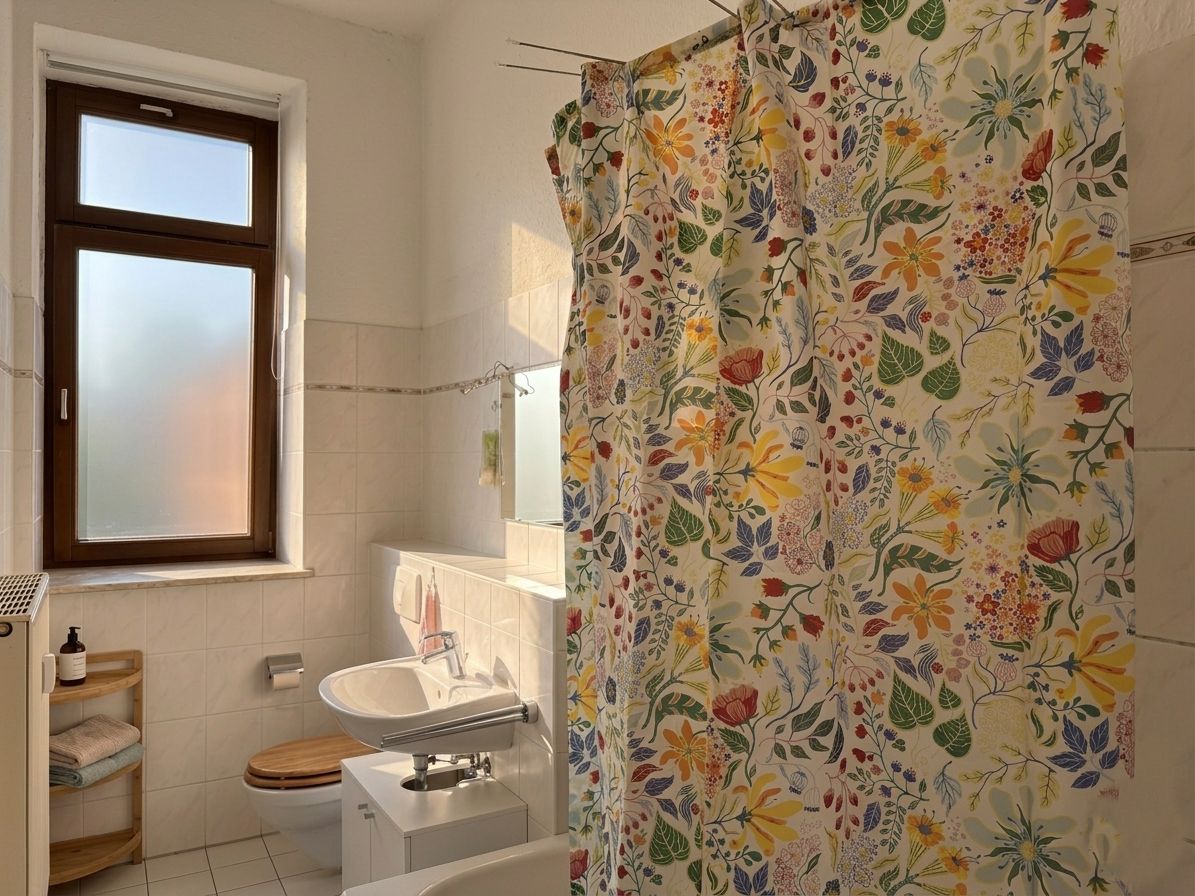 Helles Badezimmer mit Fenster, Waschbecken, Toilette und bunter Duschvorhang mit Blumenmuster.