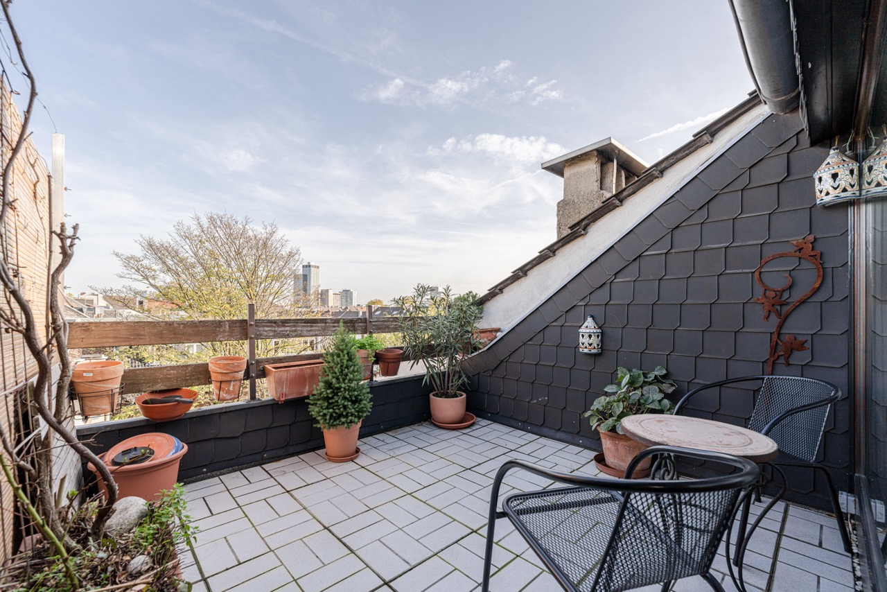 Dachterrasse mit Pflanzentöpfen, kleinem Tisch und zwei Stühlen, umgeben von Stadtblick.