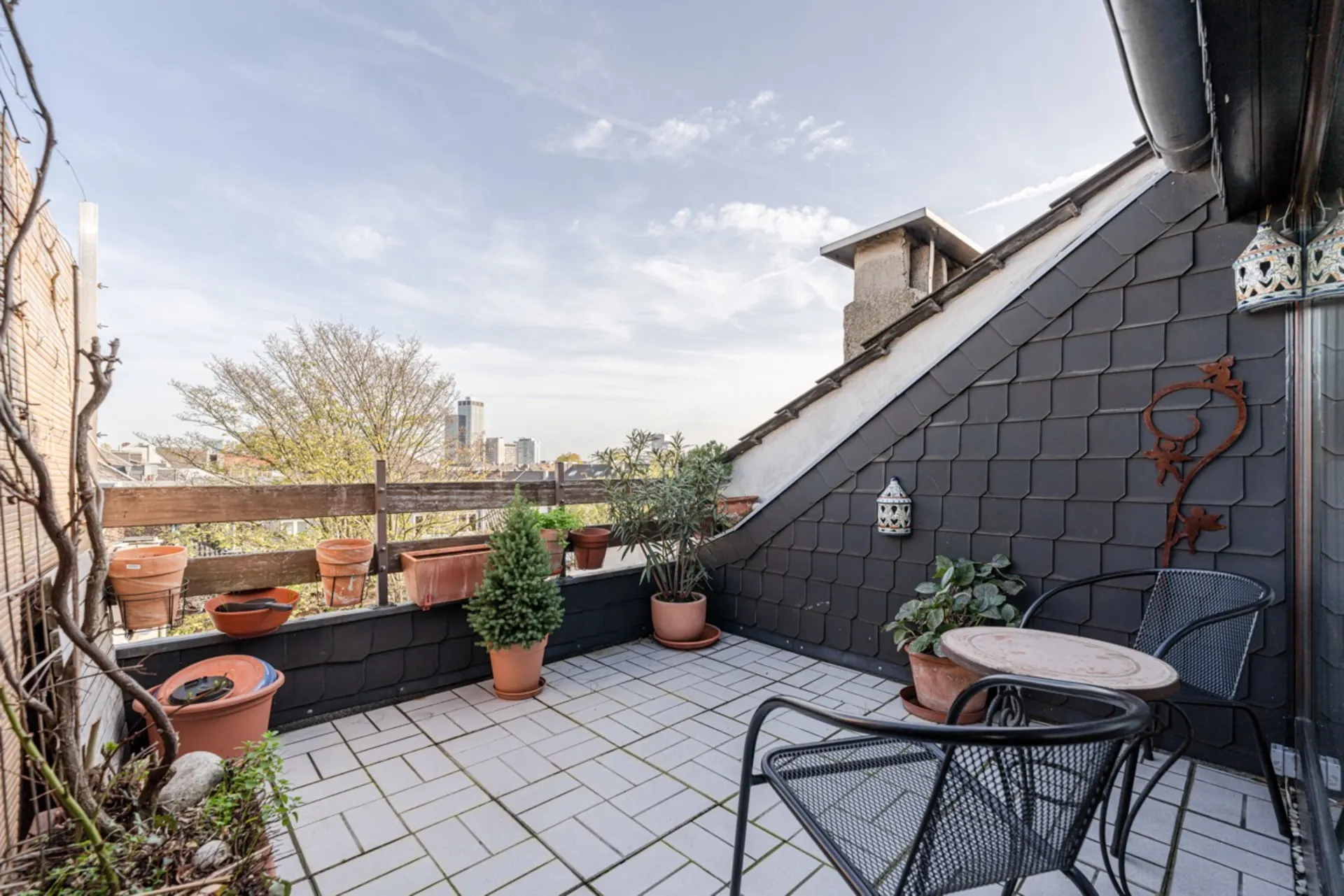 Dachterrasse mit Pflanzentöpfen, kleinem Tisch und zwei Stühlen, umgeben von Stadtblick.