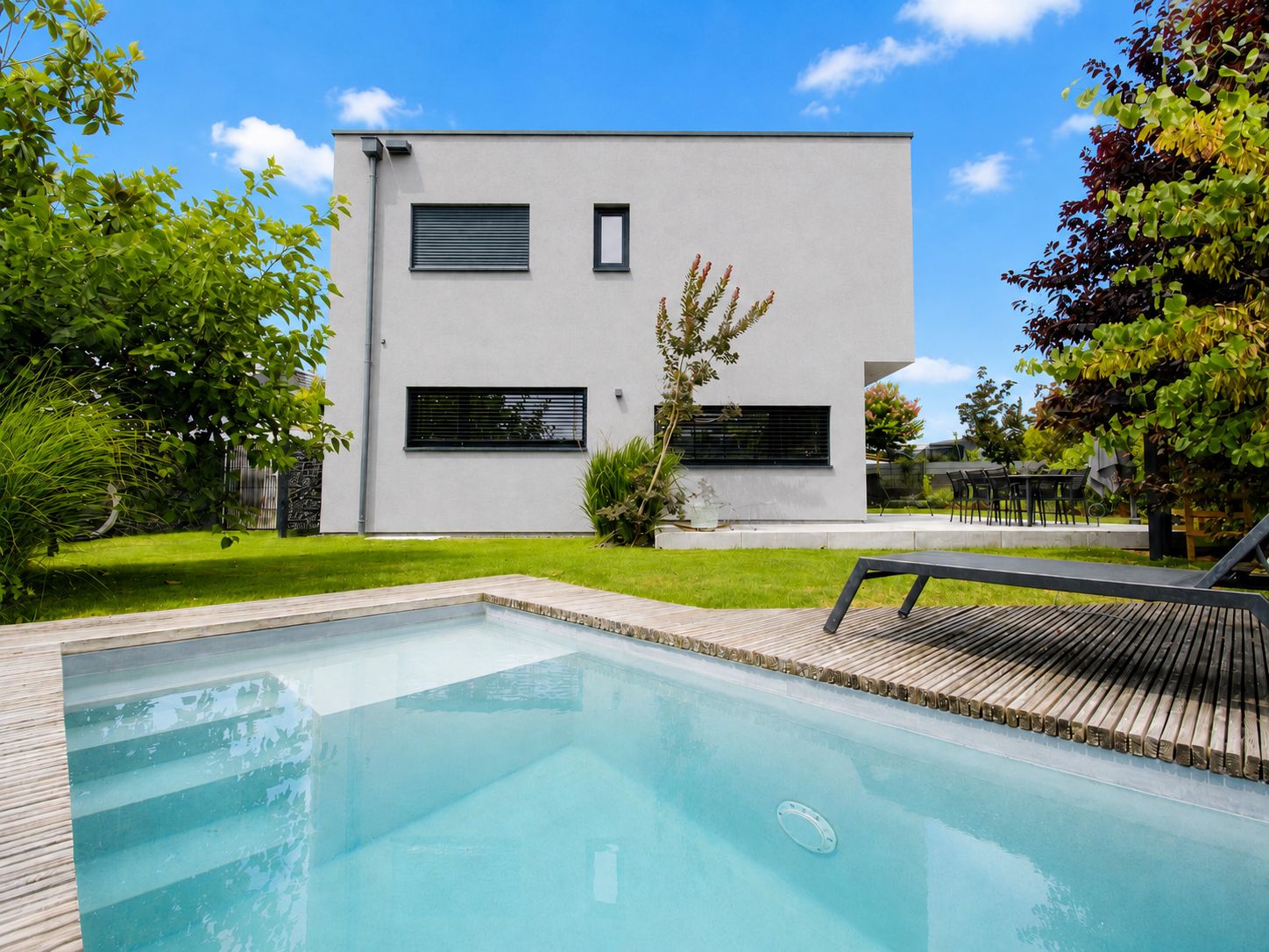 Modernes Wohnhaus mit Garten, Pool und Sonnenliege auf Holzterrasse bei blauem Himmel.