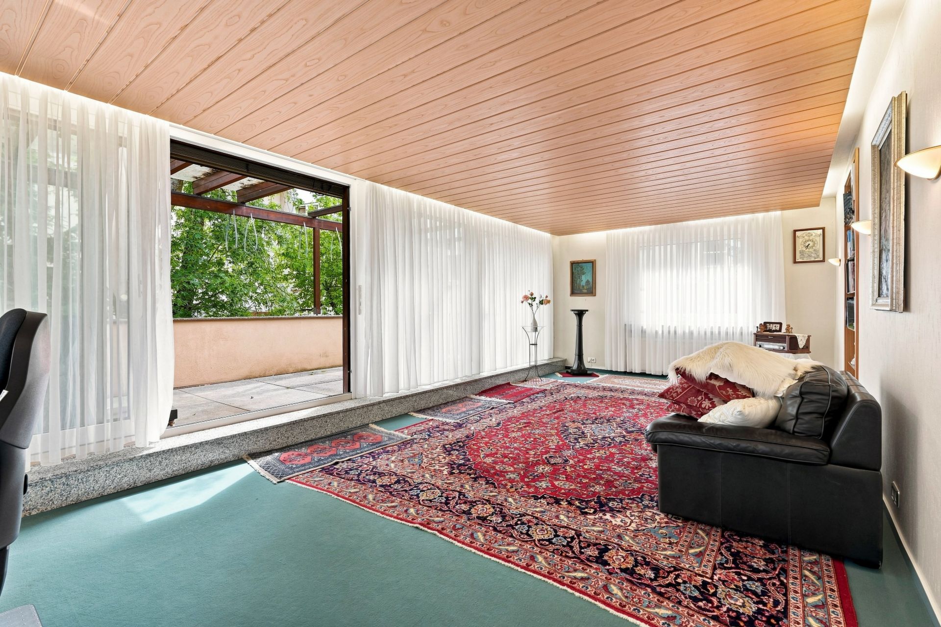 Helles Wohnzimmer mit Teppich, schwarzem Sofa und Zugang zu einer Terrasse, Vorhänge an den Fenstern.