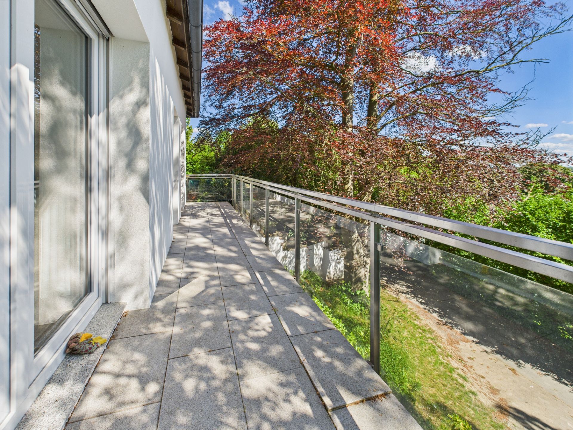 Breiter Balkon mit Glasgeländer neben einem Haus, Blick auf Bäume und blauen Himmel.