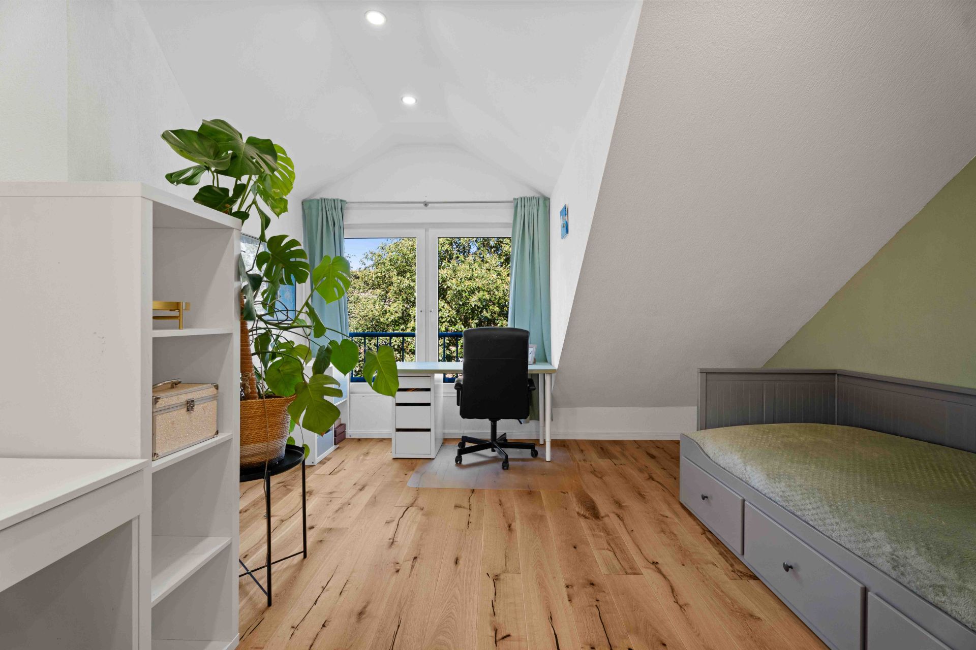 Helles Zimmer mit Holzboden, Schreibtisch am Fenster, Regal, Pflanze und Einzelbett.