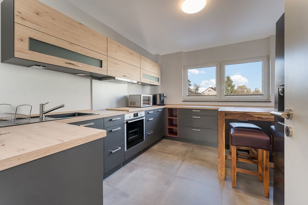 Moderne Küche mit grauen und holzfarbenen Schränken, Esstheke und zwei Fenstern.