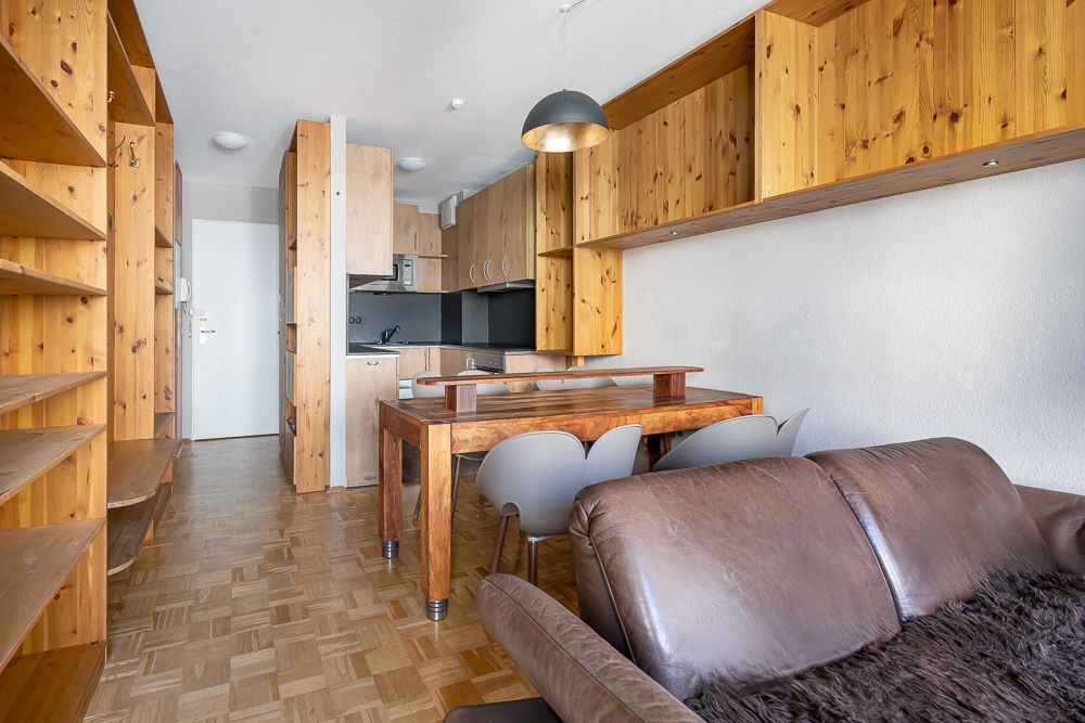 Wohnzimmer mit braunem Sofa, Holztisch, Stühlen und offener Küche mit vielen Holzregalen.