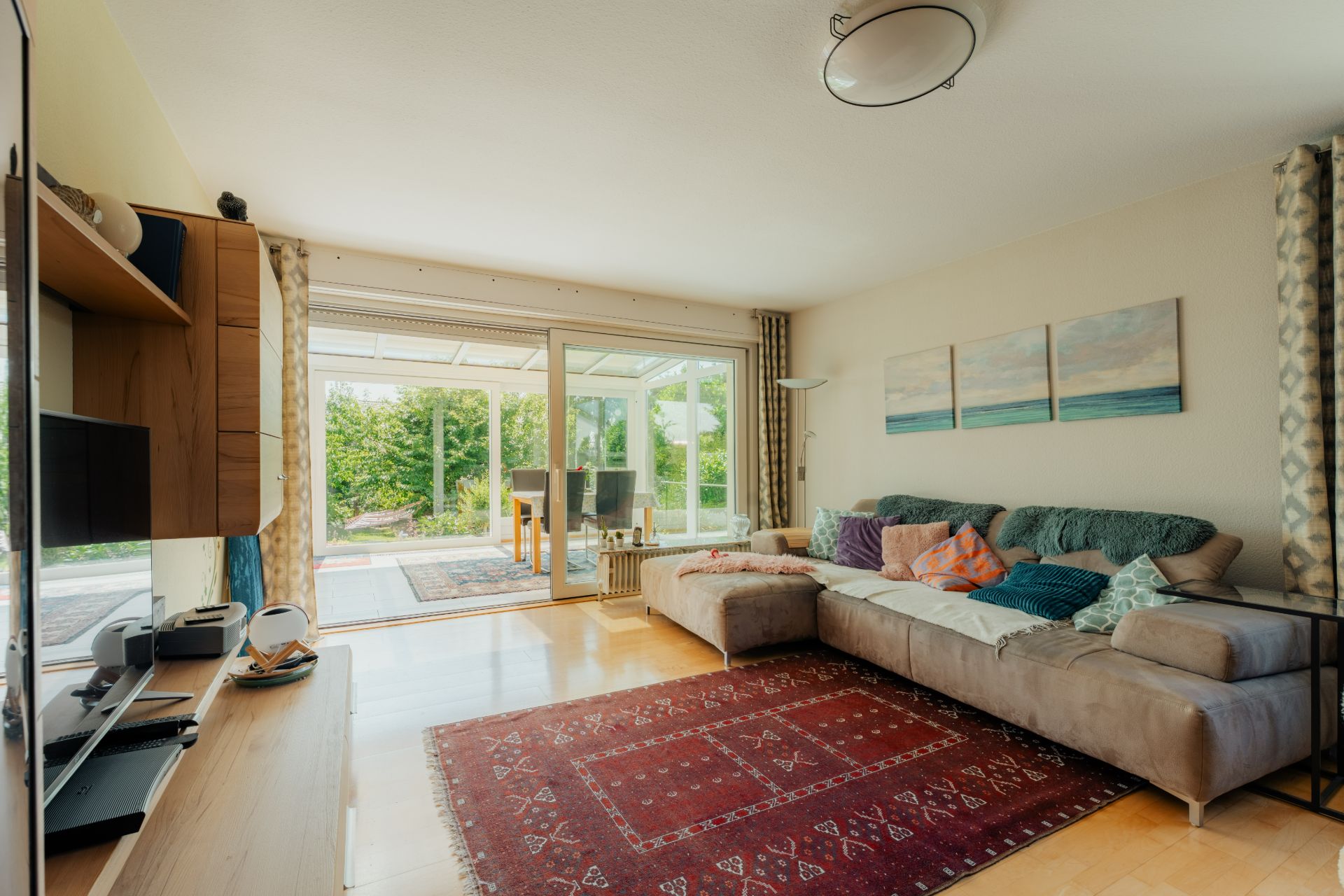 Helles Wohnzimmer mit großer Eckcouch, Teppich und Blick auf Wintergarten und Garten.