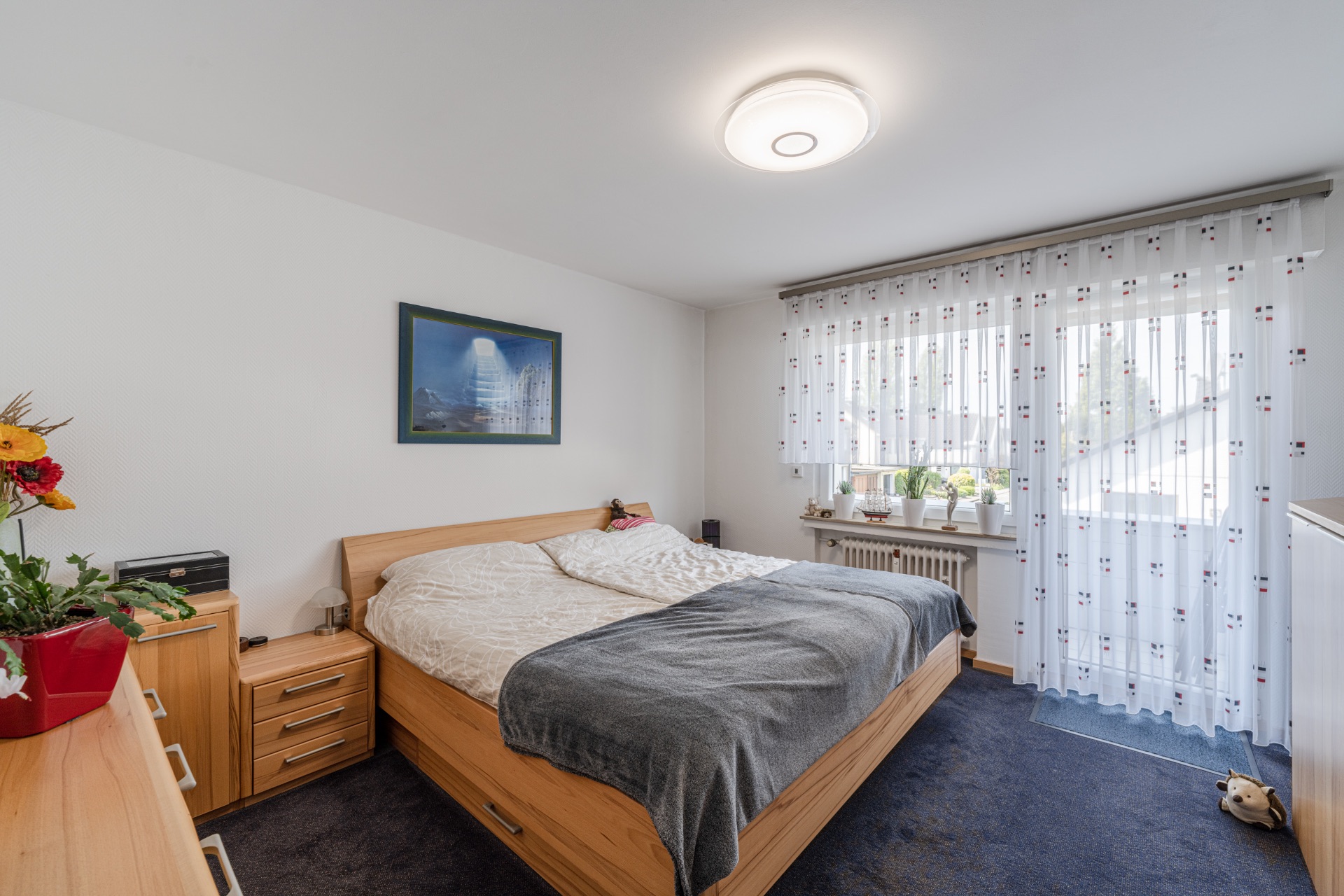 Helles Schlafzimmer mit Doppelbett, Holzmöbeln und großem Fenster mit weißen Vorhängen.