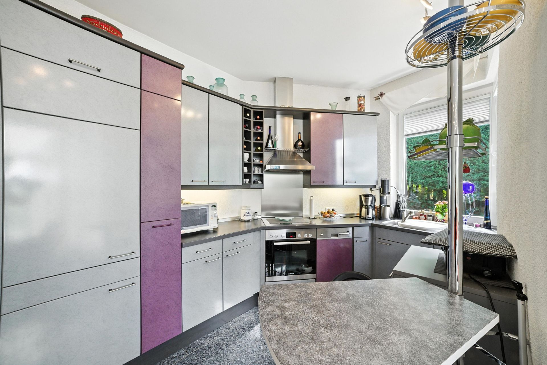 Moderne Einbauküche mit grauen und lila Fronten, Geräten und großem Fenster.