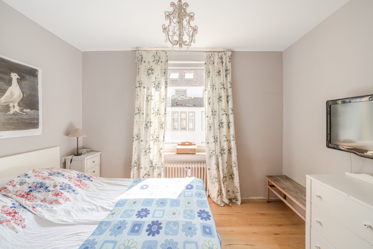 Helles Schlafzimmer mit Doppelbett, TV, Nachttisch, weißem Schrank und floralem Vorhang am Fenster.