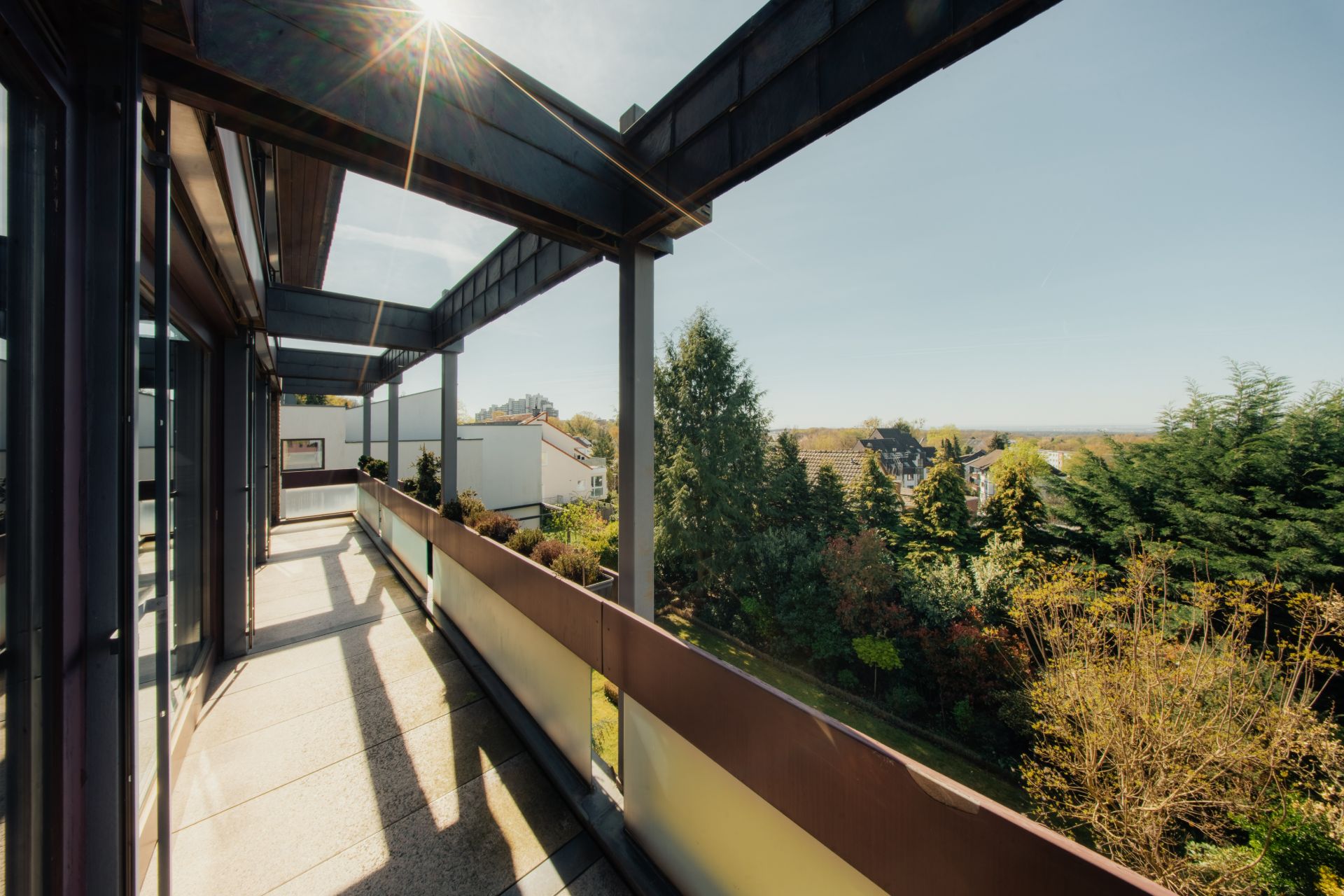 Langer Balkon mit Glasgeländer und Blick auf Bäume und benachbarte Wohnhäuser.
