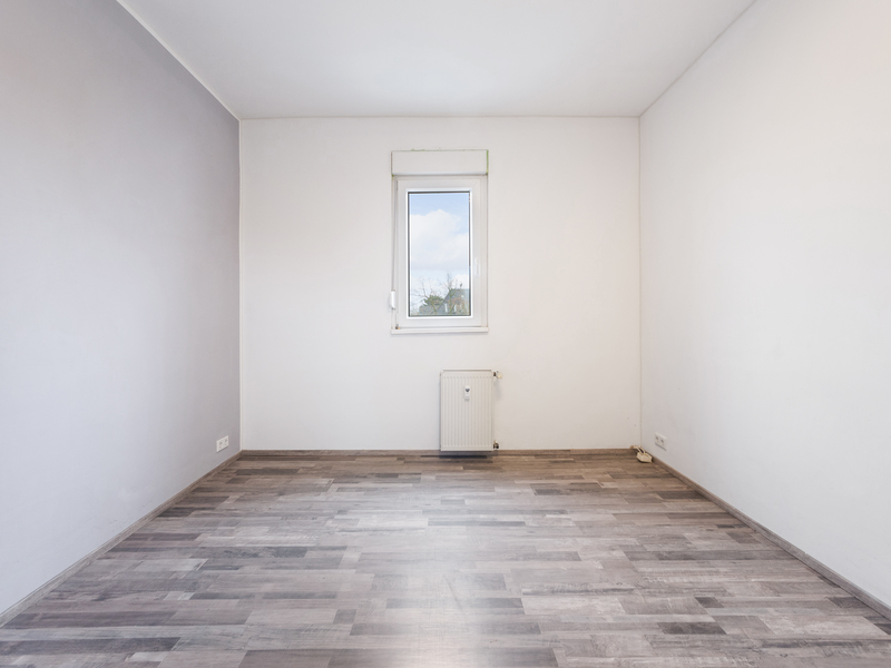 Leerer Raum mit grauem Boden, weißen Wänden, kleinem Fenster und Heizkörper.