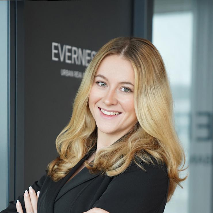 Frau mit blondem Haar in schwarzem Anzug steht lächelnd mit verschränkten Armen vor Glaswand.