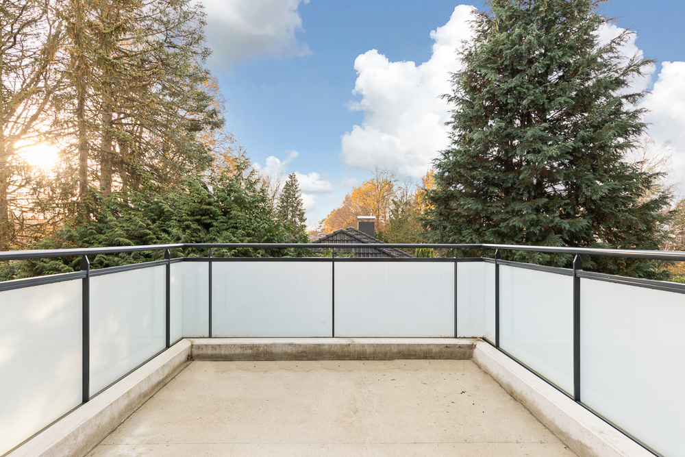Leere Terrasse mit Glasgeländer, umgeben von hohen Bäumen bei Tageslicht.