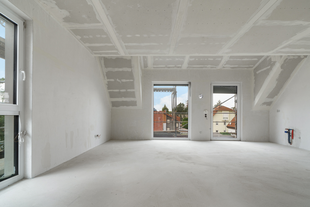 Heller, leerer Raum mit unlackierten Wänden und drei großen Fenstern, Blick auf Nachbarhäuser.