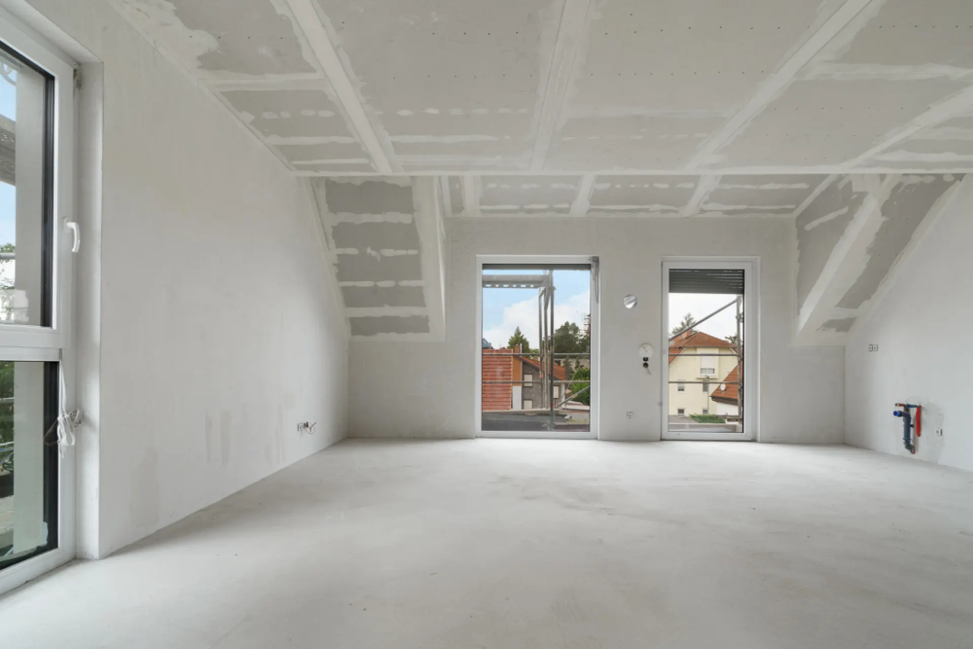 Heller, leerer Raum mit unlackierten Wänden und drei großen Fenstern, Blick auf Nachbarhäuser.