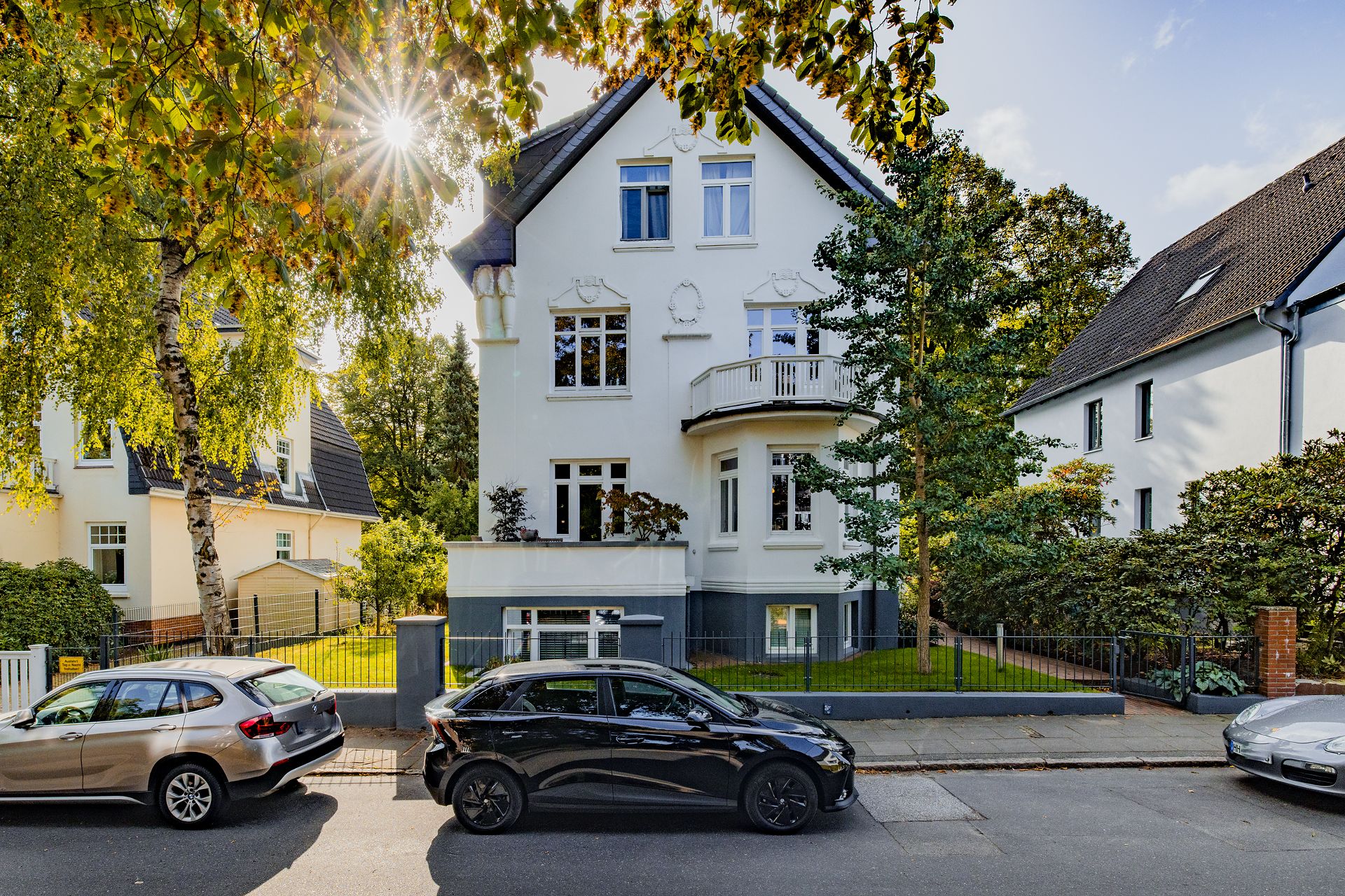 Weiße Villa mit Balkon und Garten, vorne geparkte Autos, Bäume im Vordergrund.
