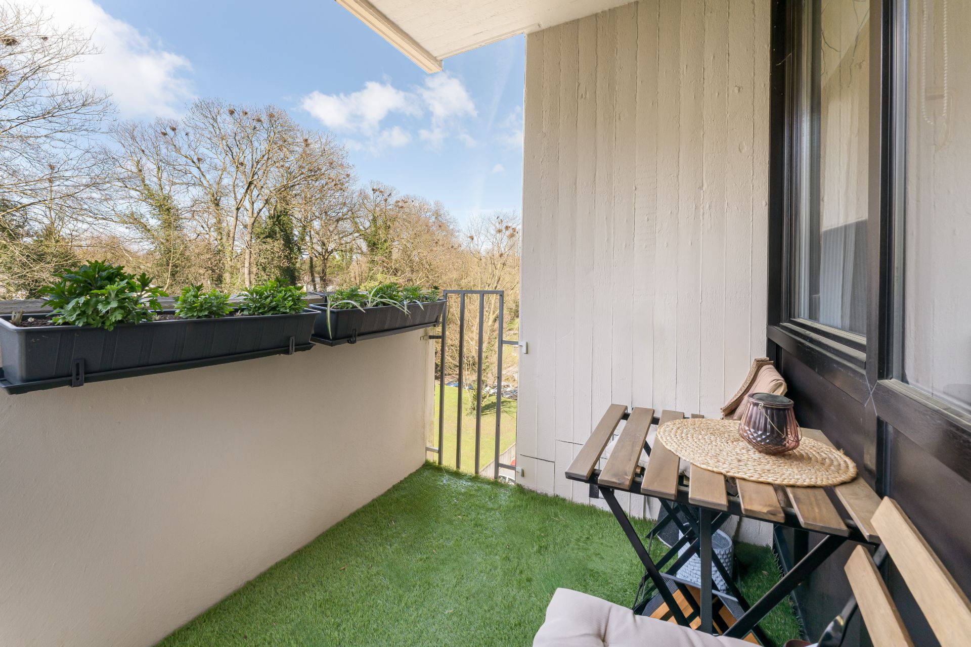 Kleiner Balkon mit Kunstrasen, Holztisch, Stühlen und Blumenkästen am Geländer.