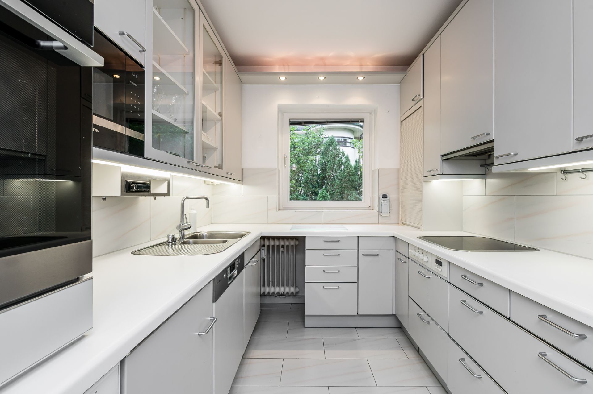 Moderne, helle Einbauküche mit weißen Schränken, Geräten und Fenster mit Blick ins Grüne.