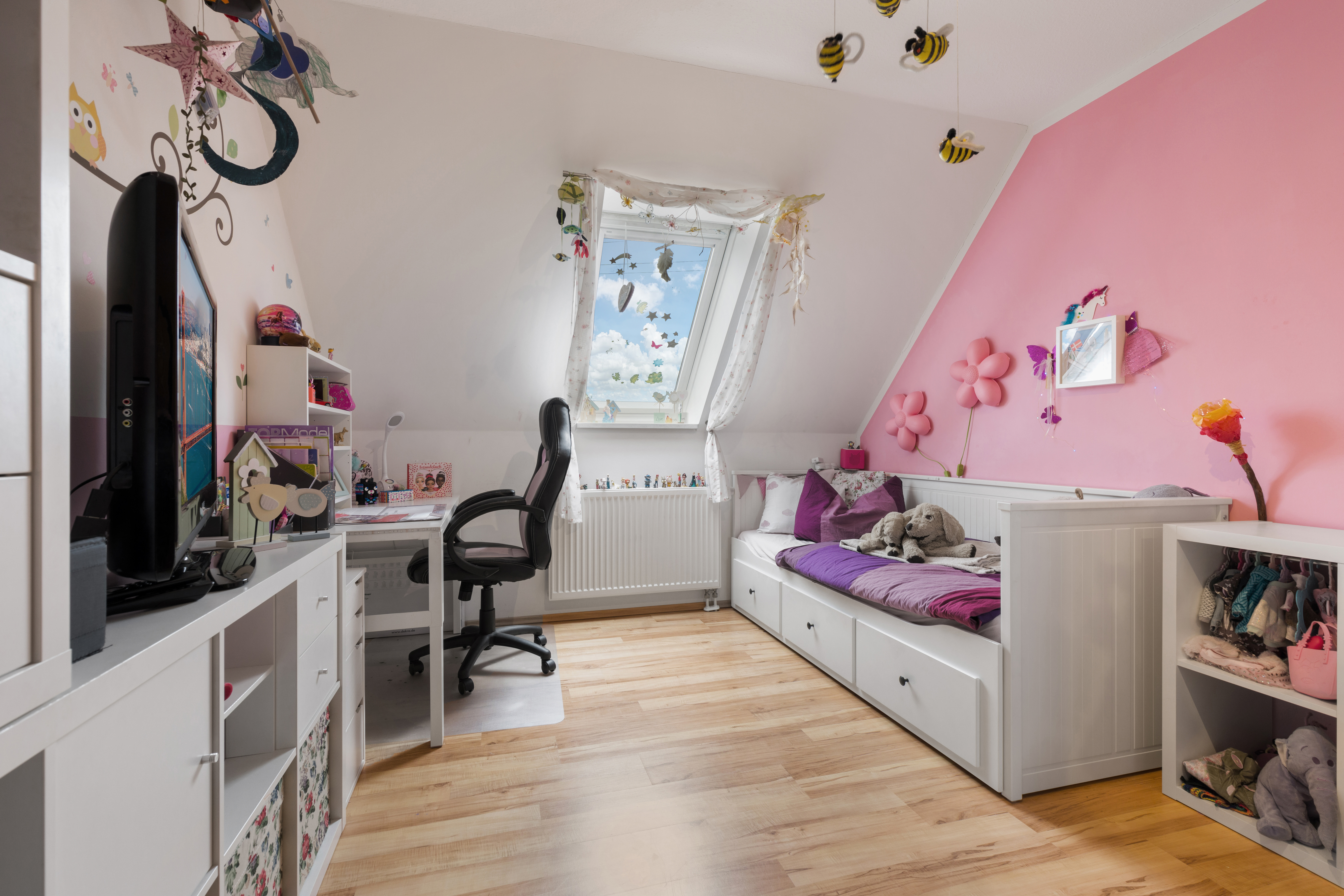 Kinderzimmer mit Schreibtisch, Regal und Bett unter einem Dachfenster, pinke Wanddekoration.