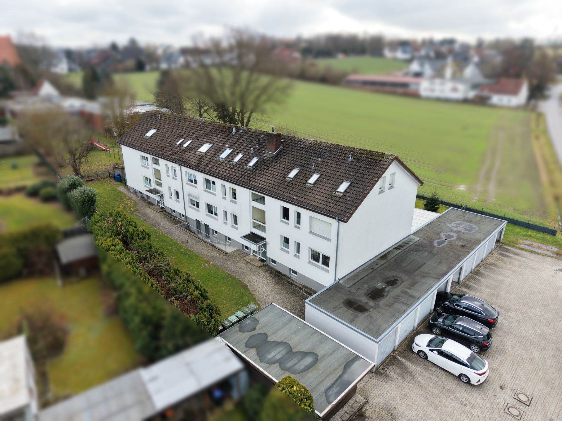 Mehrfamilienhaus mit Satteldach, Garagen und geparkten Autos, daneben eine Grünfläche.