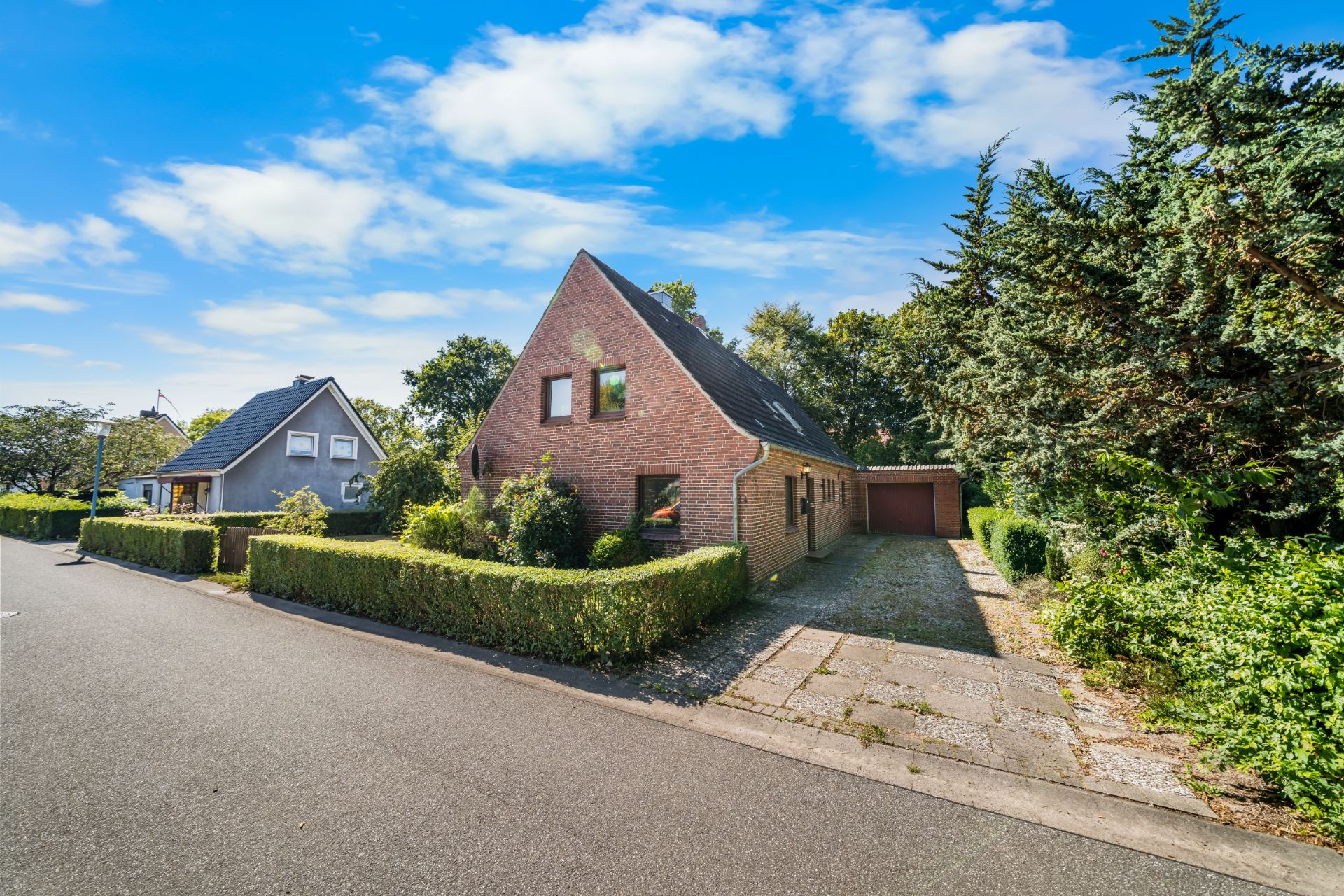 Backsteinhaus mit Spitzdach, Einfahrt und Garage, umgeben von Hecken und Bäumen an einer ruhigen Straße.