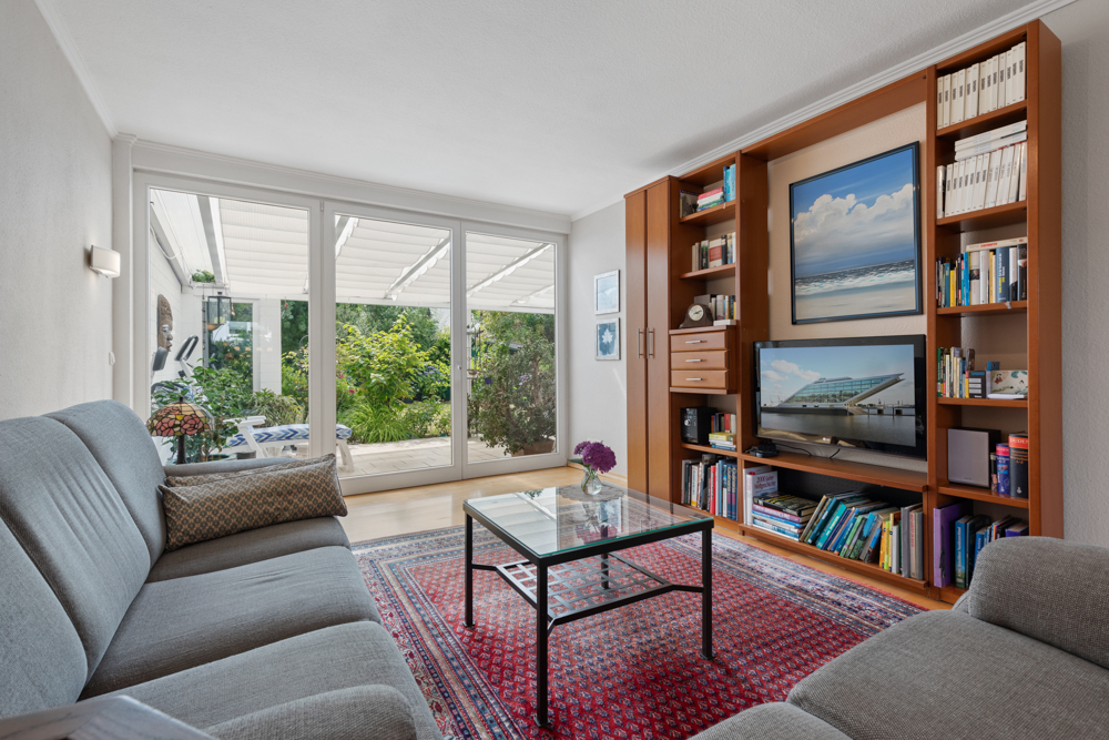 Helles Wohnzimmer mit grauen Sofas, Glastisch, Bücherregal und großen Fenstern zum Garten.