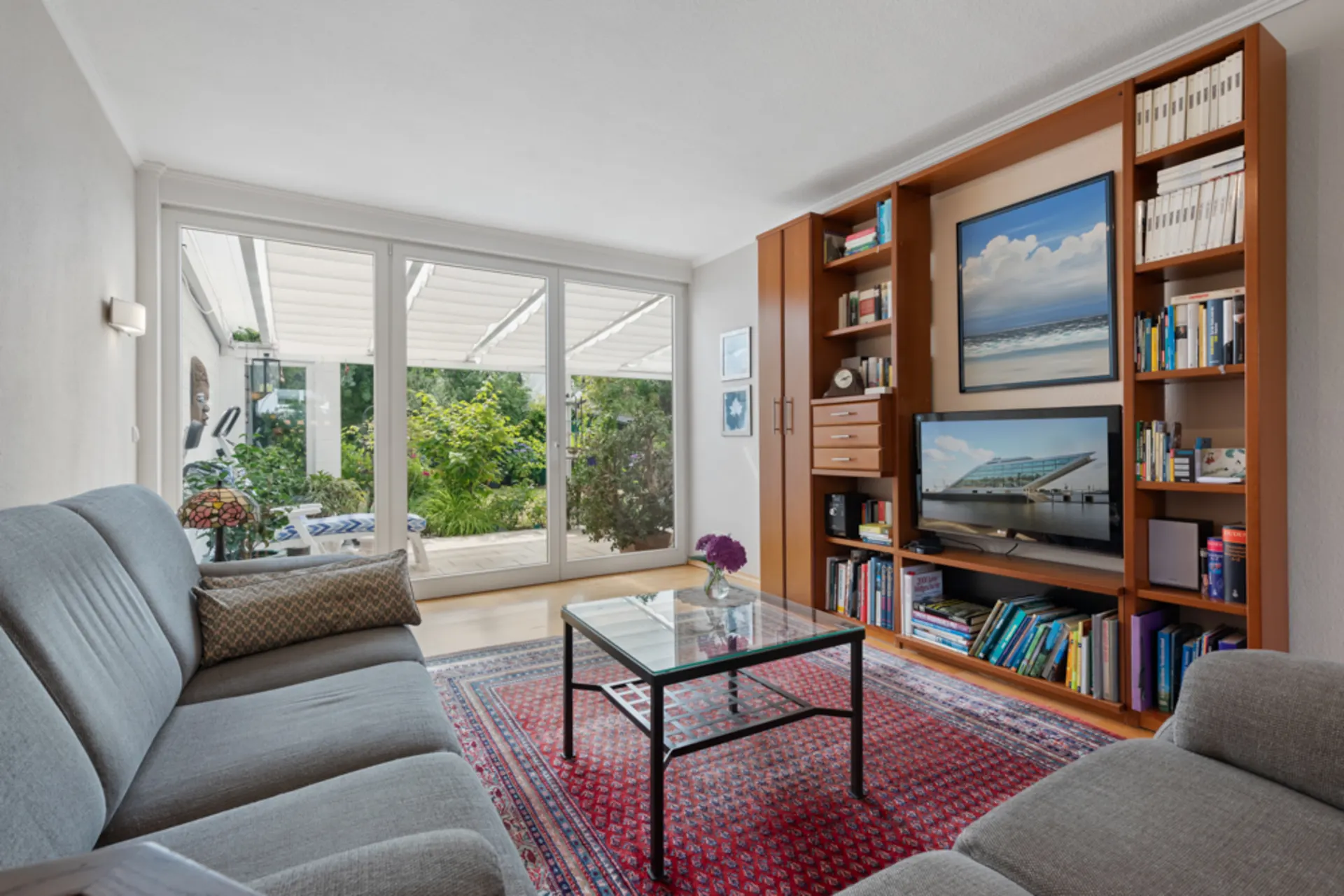 Helles Wohnzimmer mit grauen Sofas, Glastisch, Bücherregal und großen Fenstern zum Garten.