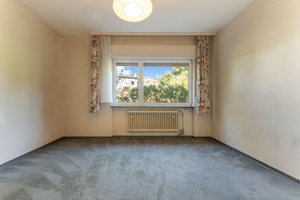 Leeres Zimmer mit Teppichboden, großem Fenster, Heizung und gemusterten Vorhängen.