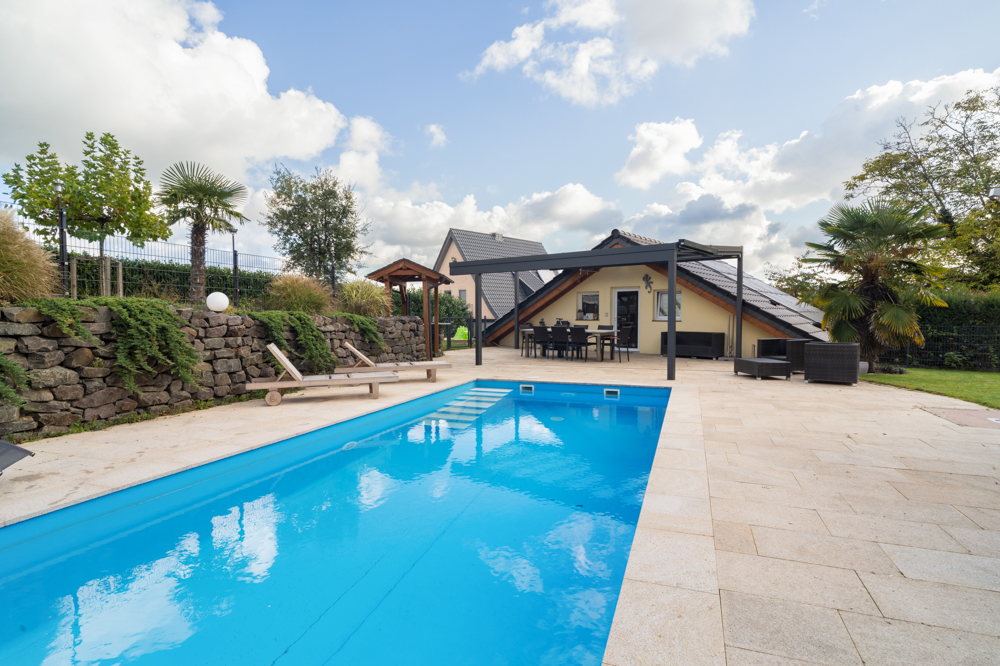 Garten mit Pool, Sonnenliegen, Palmen und moderner Terrasse vor einem Einfamilienhaus.