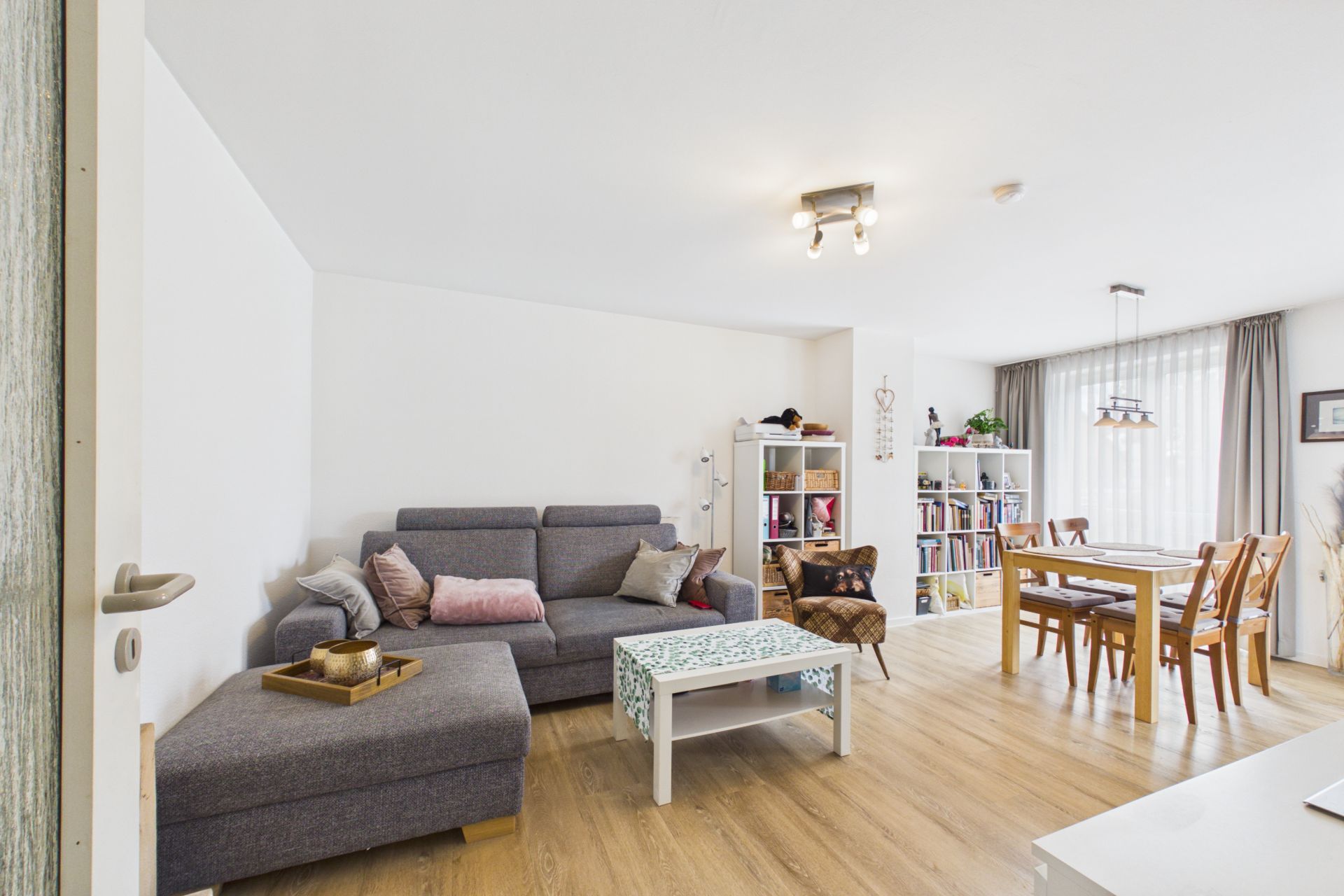 Helles Wohnzimmer mit grauem Sofa, Couchtisch, Bücherregalen und Esstisch mit Stühlen.