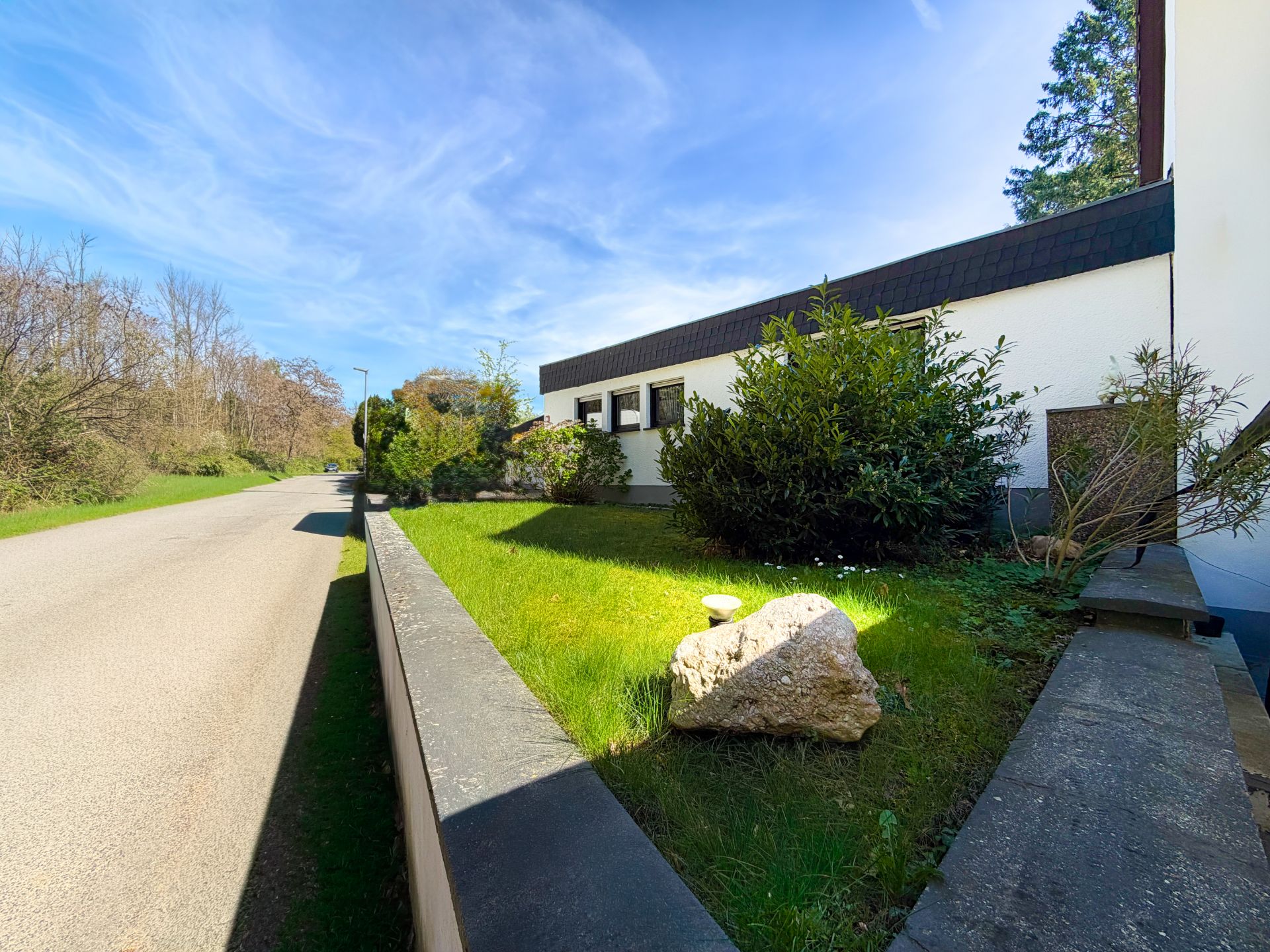 Gepflegter Vorgarten mit Strauch, Rasen und Stein neben einem weißen Wohnhaus an einer ruhigen Straße.