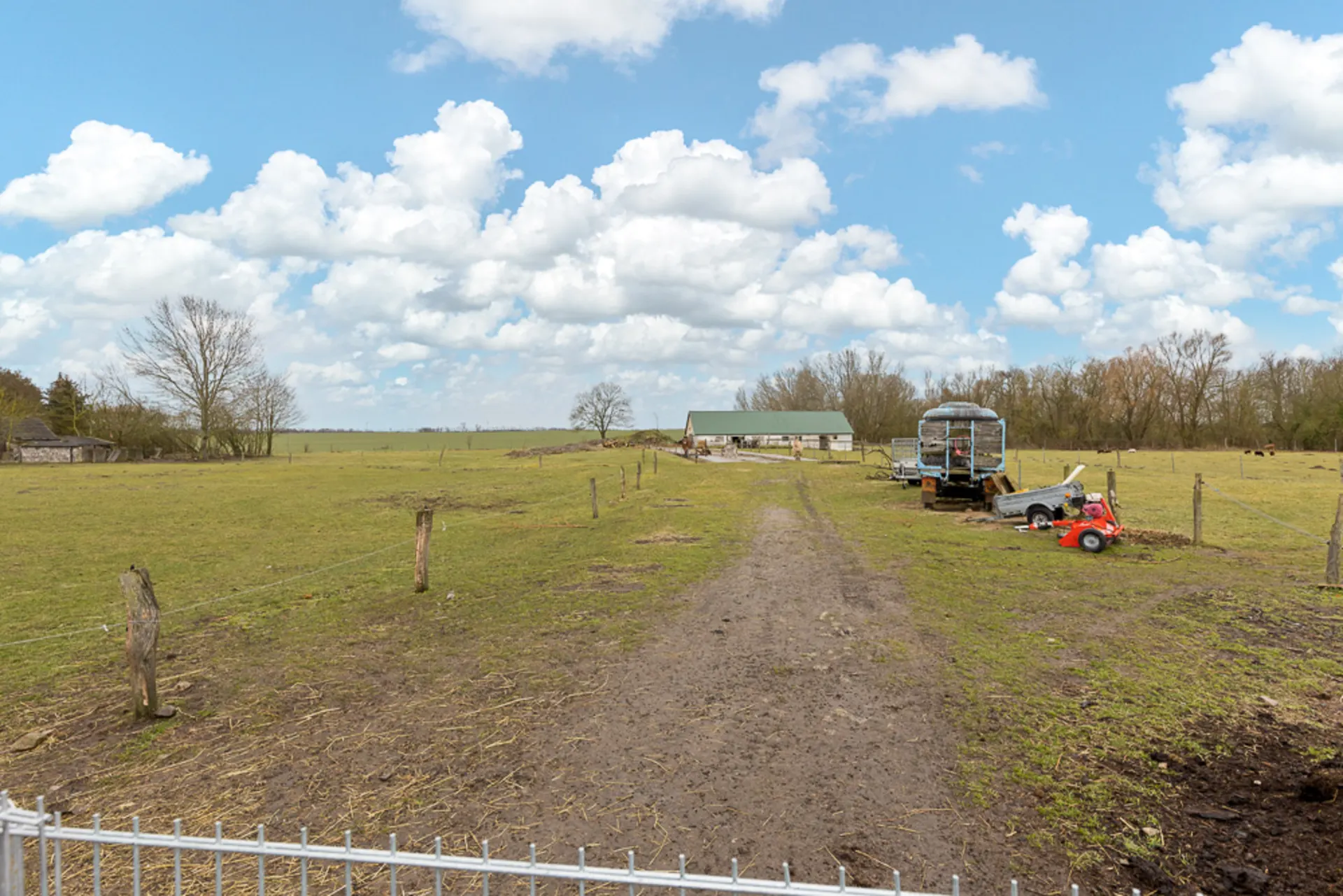 Breiter Feldweg mit Zaun, Anhängern und Traktor auf grüner Wiese mit Bäumen und Haus im Hintergrund.
