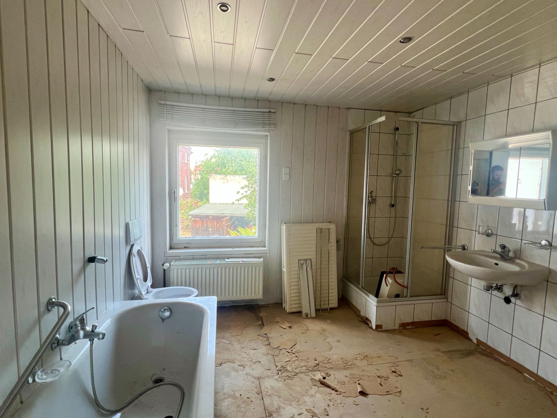 Helles Badezimmer mit Badewanne, Dusche, Waschbecken und Fenster, Boden teilweise beschädigt.