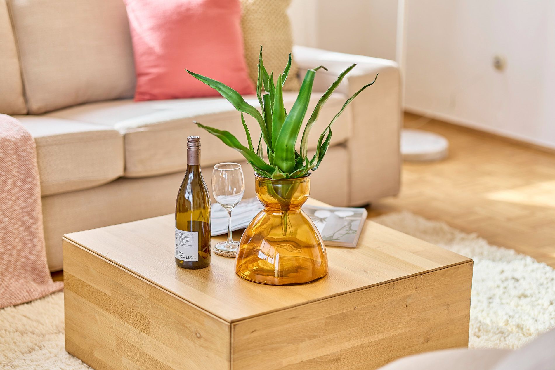 Holztisch mit Pflanze in Vase, Weinflasche und Weinglas vor hellen Sofas und Kissen.