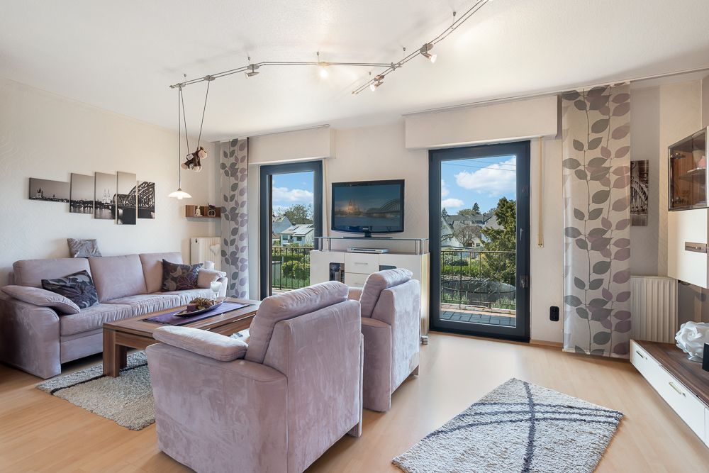 Helles Wohnzimmer mit Sofa, zwei Sesseln, Couchtisch, TV und großen Fenstern zum Balkon.