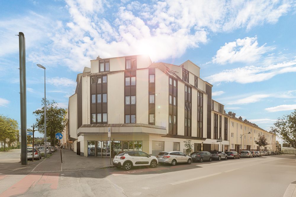 Mehrstöckiges Wohnhaus an einer Straßenecke mit geparkten Autos und blauem Himmel.