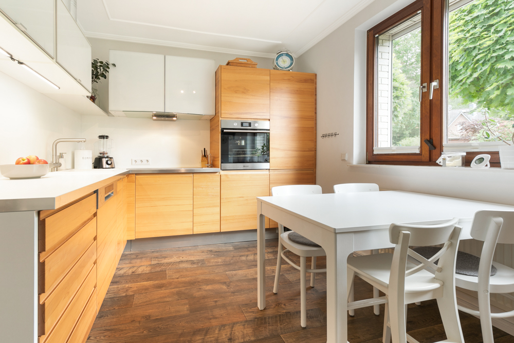 Moderne Küche mit Holzmöbeln, weißem Esstisch und vier Stühlen vor einem Fenster.