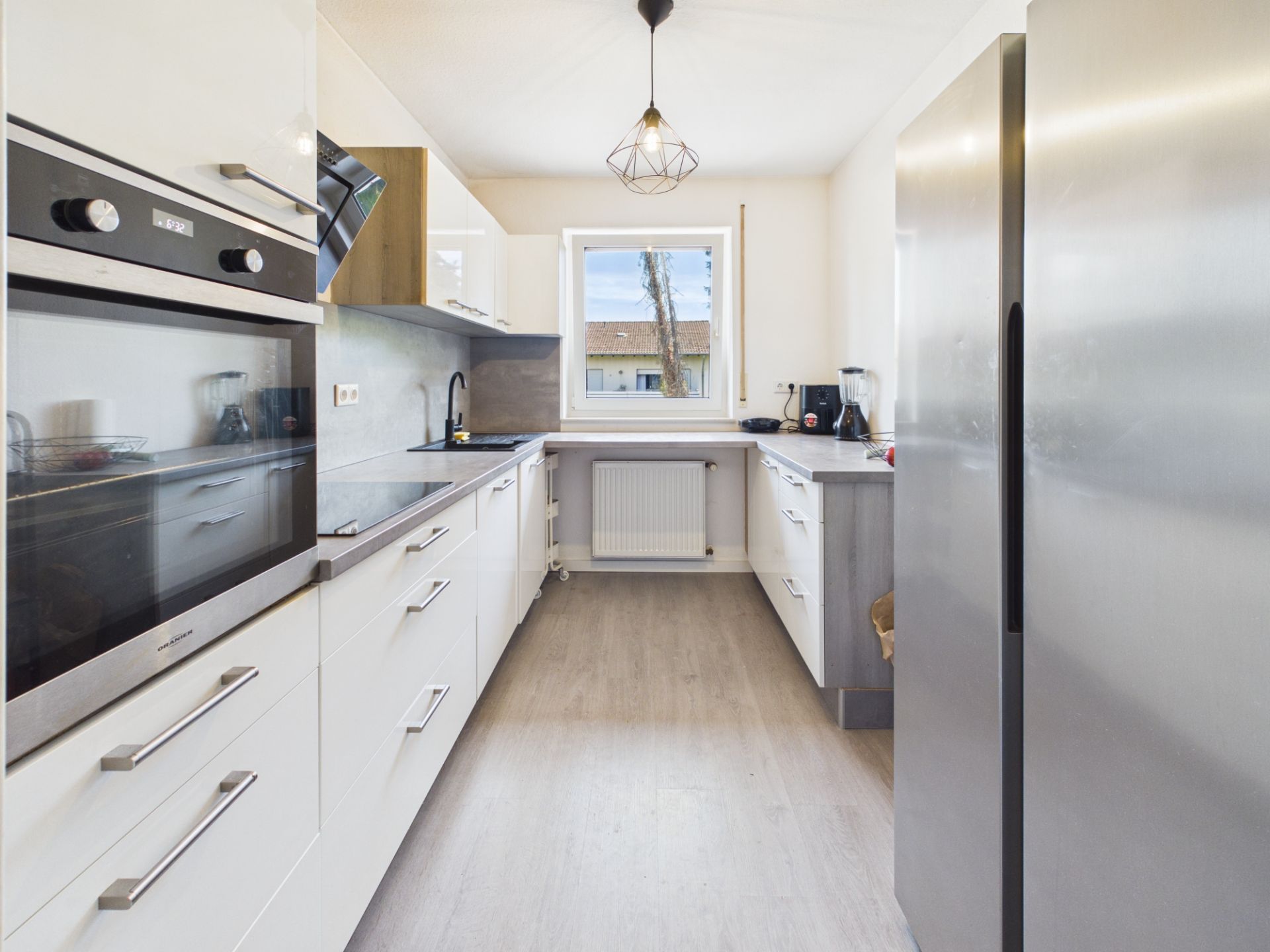Moderne, helle Küche mit weißen Schränken, großem Kühlschrank und Fenster mit Blick nach draußen.