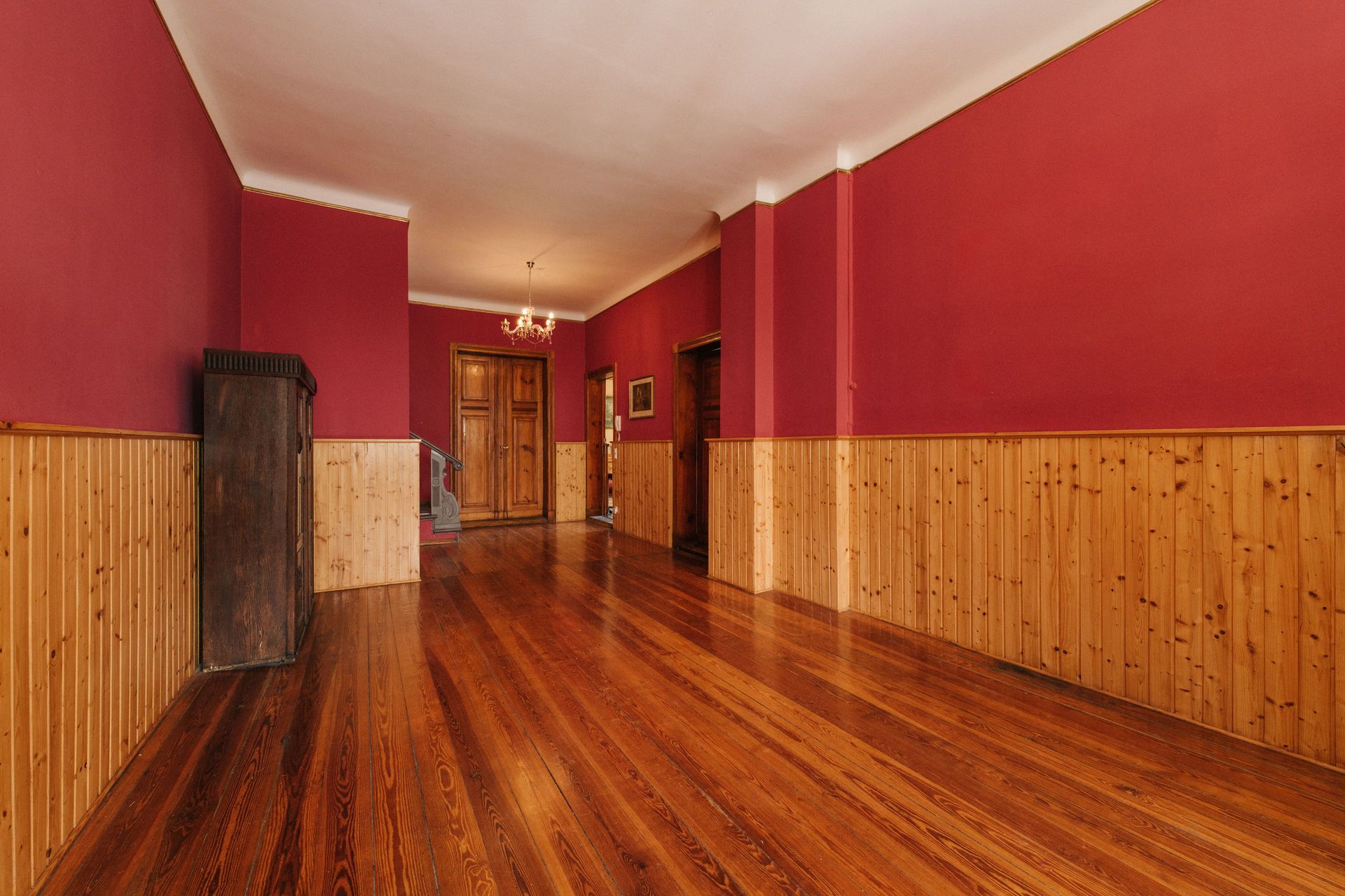Langer Flur mit Holzboden, roten Wänden und Holzvertäfelung, Kronleuchter an der Decke.